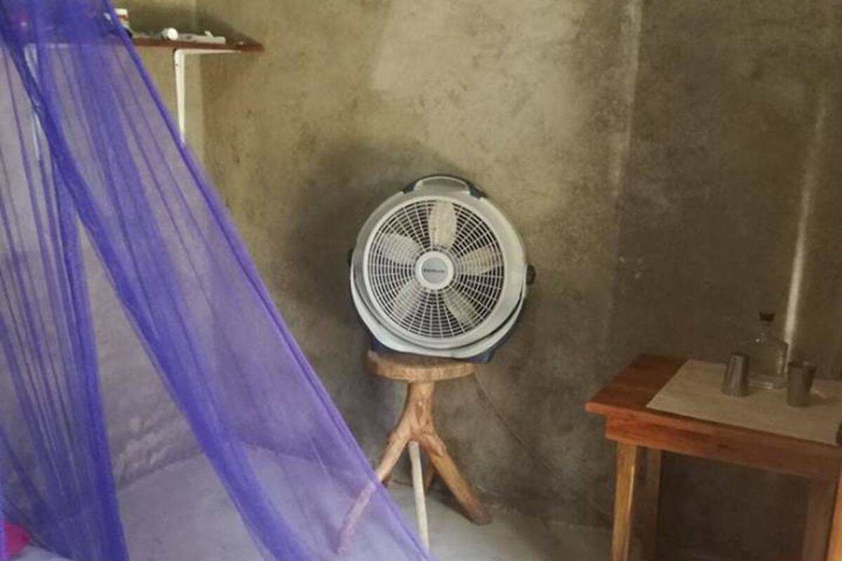 A room with a bed under a blue canopy, a small wooden table, and a standing fan against a concrete wall.