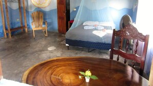 Un dormitorio con una cama cubierta por una mosquitera, una silla de madera, una mesa redonda de madera con una flor, y una pared con un diseño circular grande.