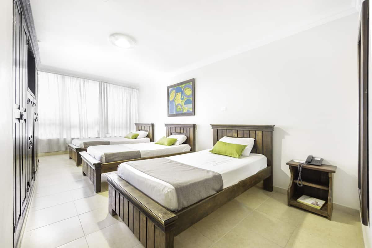 Quarto de hotel com três camas de solteiro, um guarda-roupa de madeira, mesa de cabeceira com telefone e um quadro na parede.