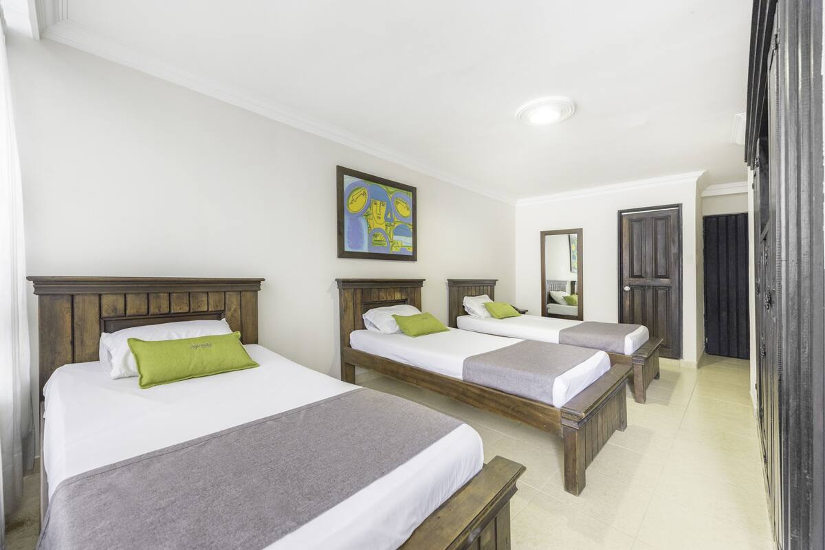 Quarto de hotel com três camas individuais, cada uma com cabeceira de madeira e roupa de cama branca. Há um quadro na parede acima das camas, um espelho na parede à direita e uma porta à direita do espelho.