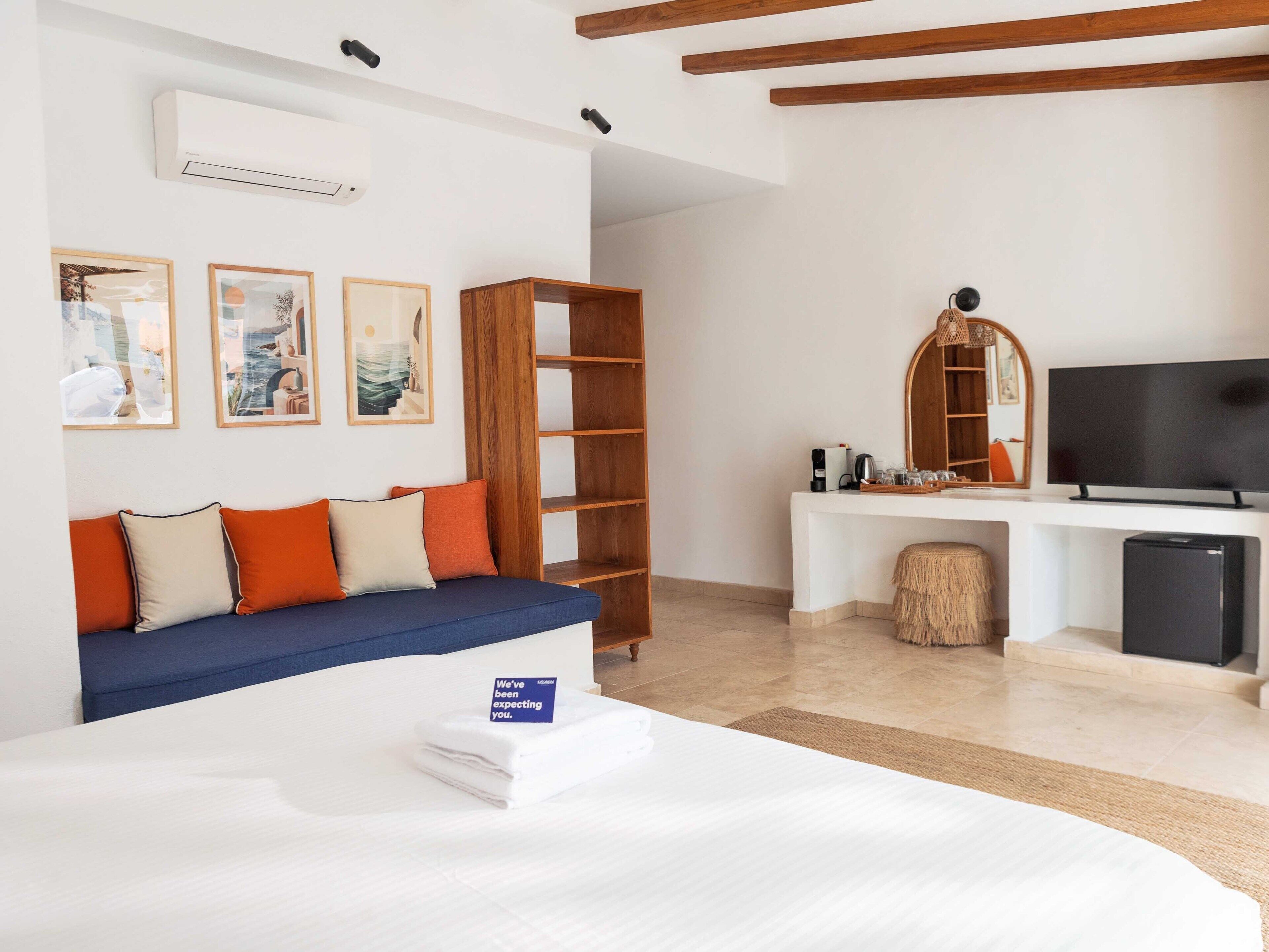 A hotel room with a bed, a sofa, a desk with a TV, a bookshelf, and artwork on the wall.