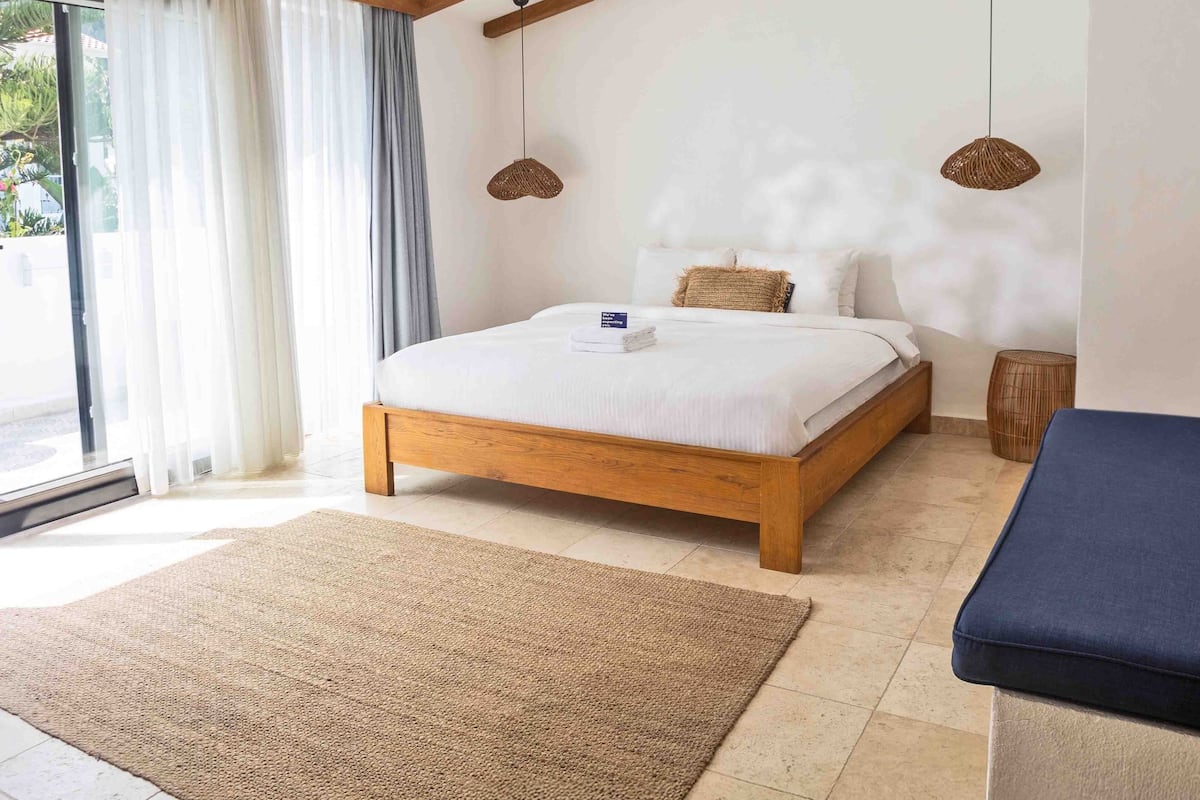 A bedroom with a wooden bed, a large window with curtains, a woven rug, and two hanging light fixtures.