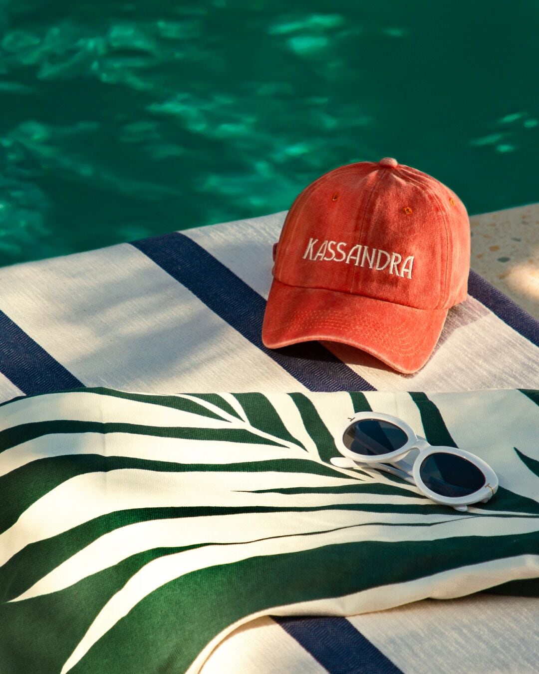 A red cap with "KASSANDRA" embroidered on it, sunglasses, and a striped towel by a pool.