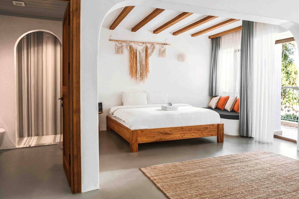 A bedroom with a wooden bed, a wall-mounted macrame piece, and a view of the outdoors.