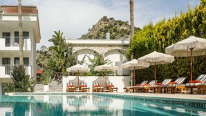 Un área junto a la piscina con sillas de descanso y sombrillas, una pared de piedra con arcos y un edificio con balcones.
