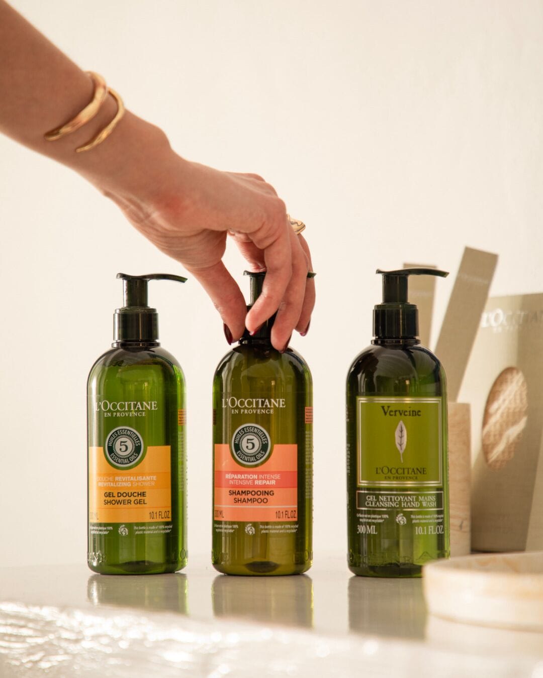 Three bottles of L'OCCITANE products on a table with a hand reaching for the middle bottle.