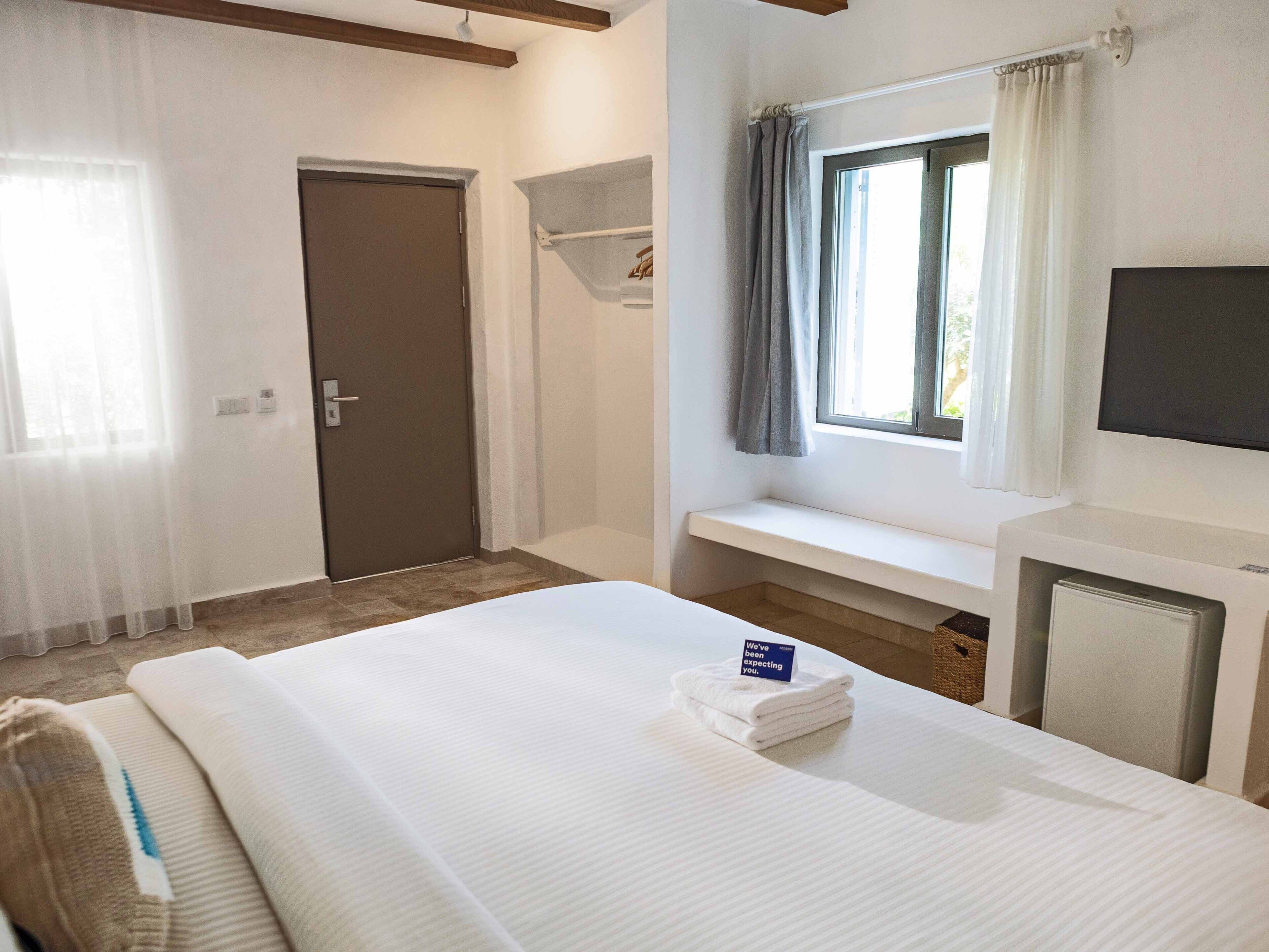Habitación de hotel con cama, televisión, ventana con cortinas, puerta y una pequeña estantería.