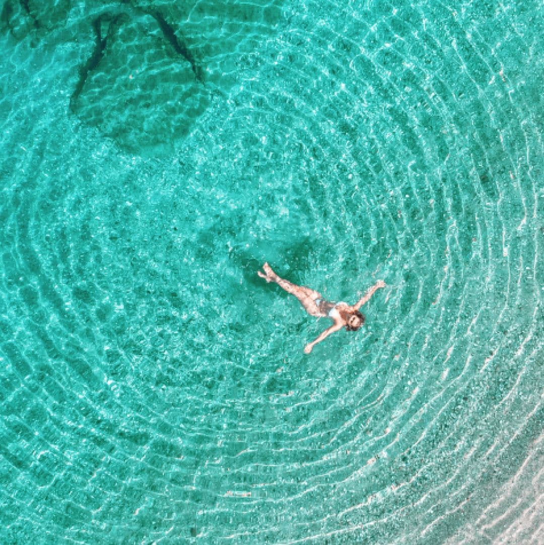 An aerial view of a person swimming in clear turquoise water with visible ripples.