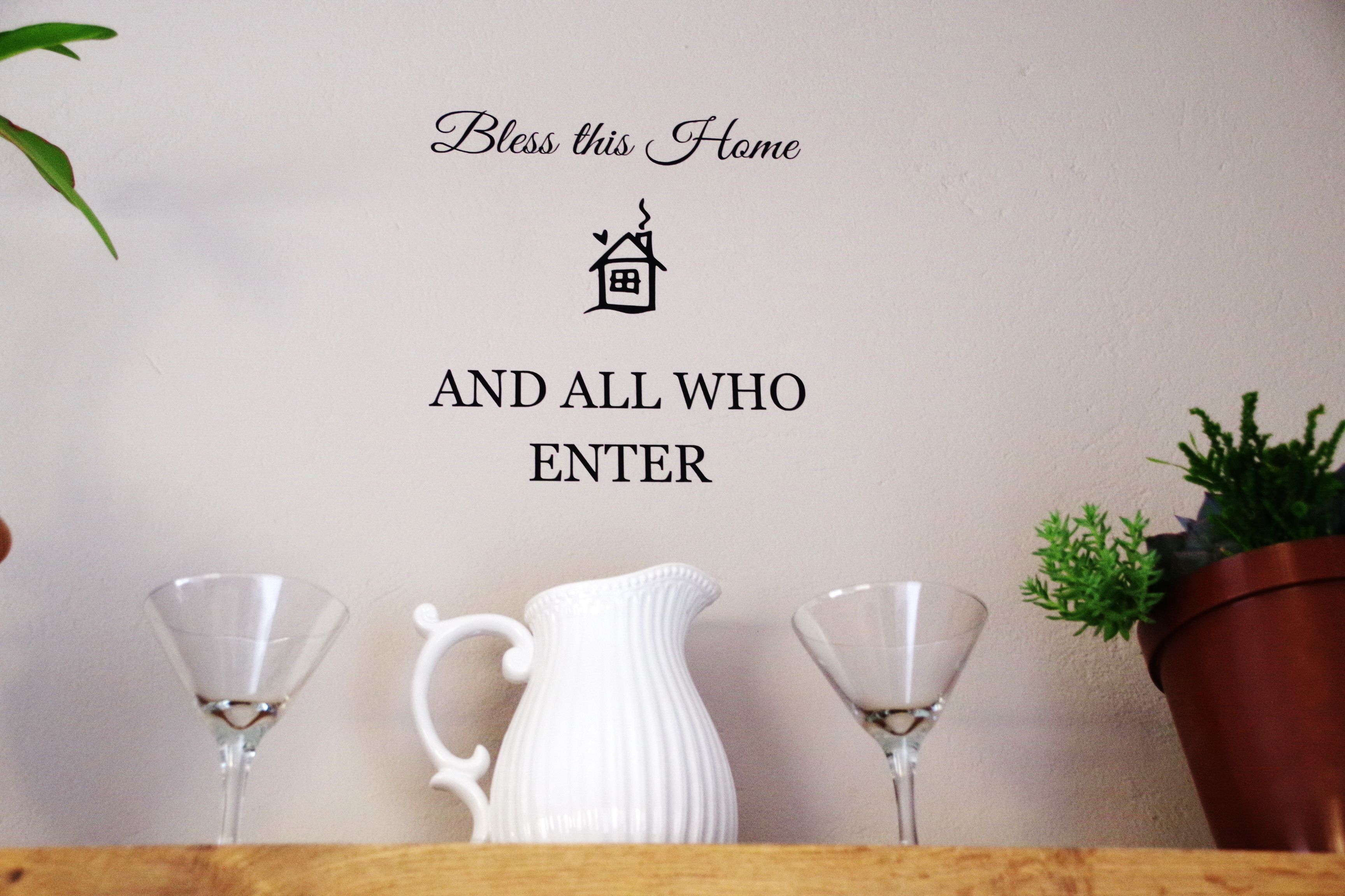 En væg med teksten "Bless this Home AND ALL WHO ENTER" og et lille husikon. Der er to martini-glas og en hvid kande på en træoverflade.