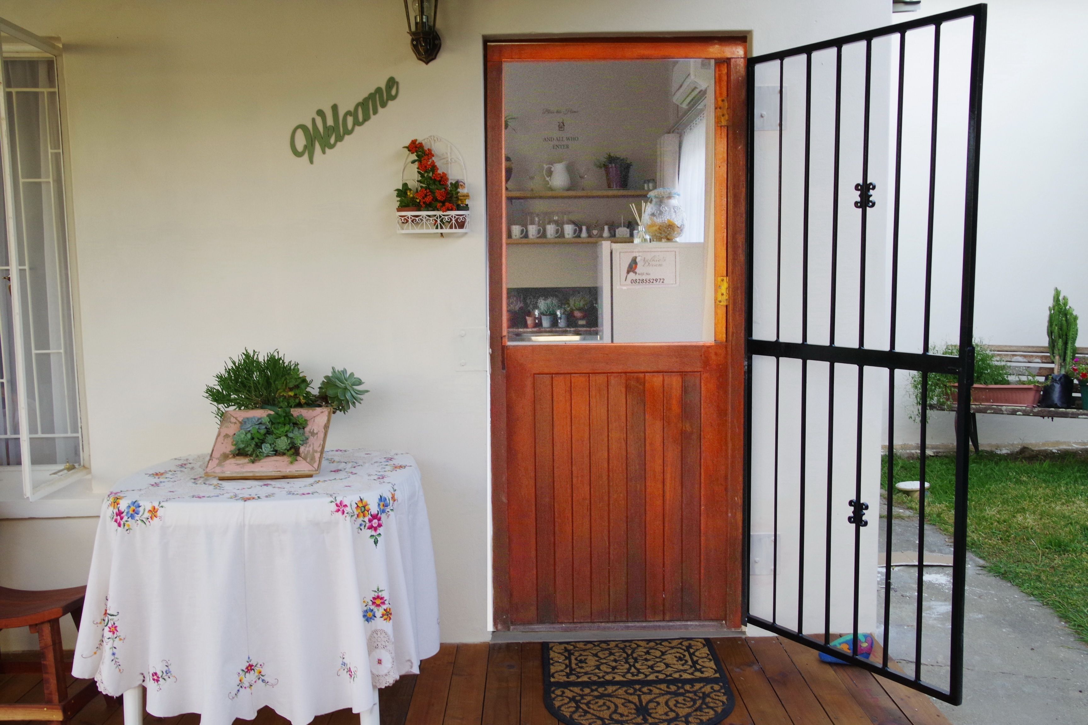 A wooden door with a black metal gate, a welcome sign, a table with a floral tablecloth, and a small potted plant.