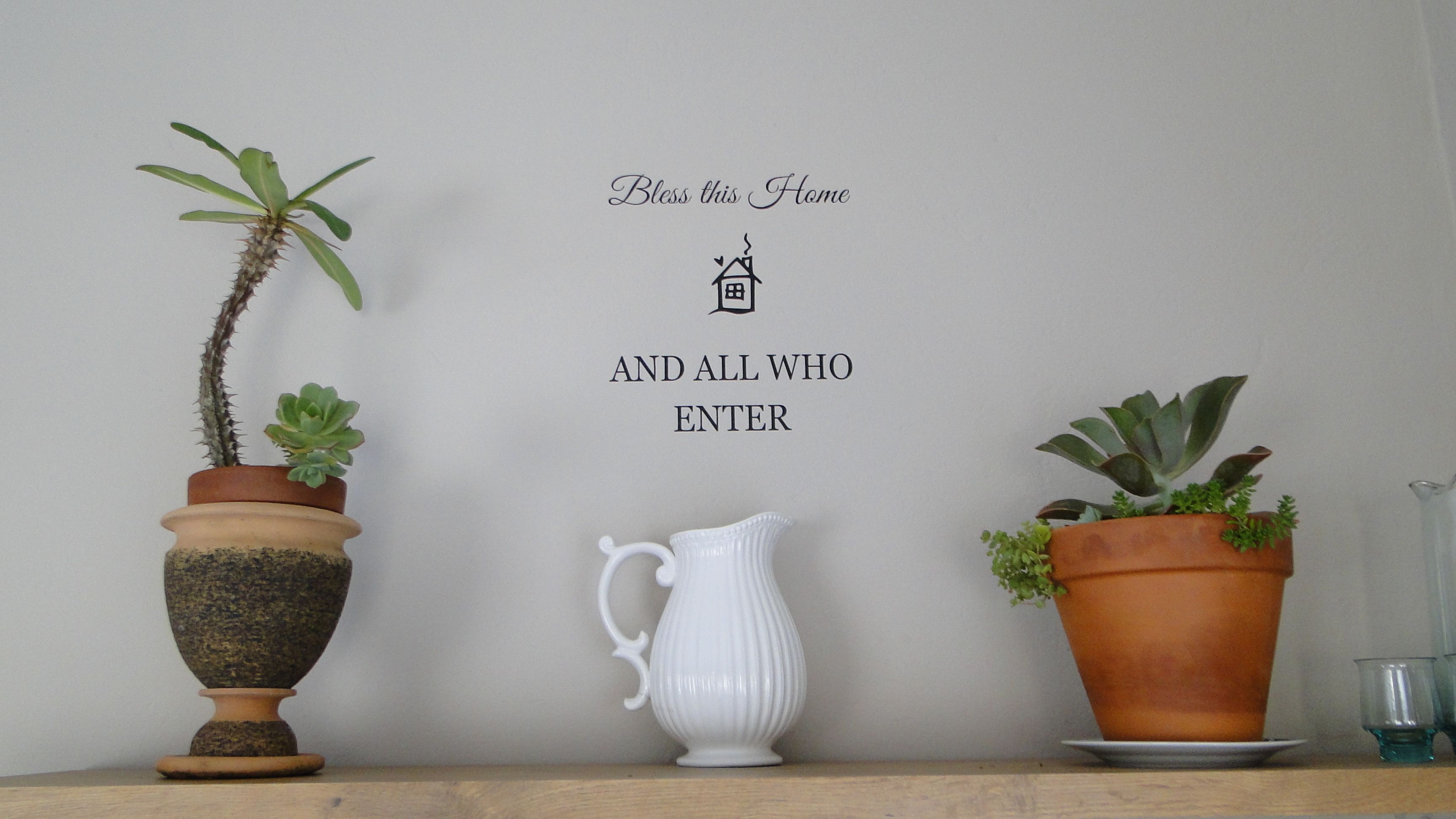 A wall with a sign that reads "Bless this Home AND ALL WHO ENTER" and three potted plants on a shelf.