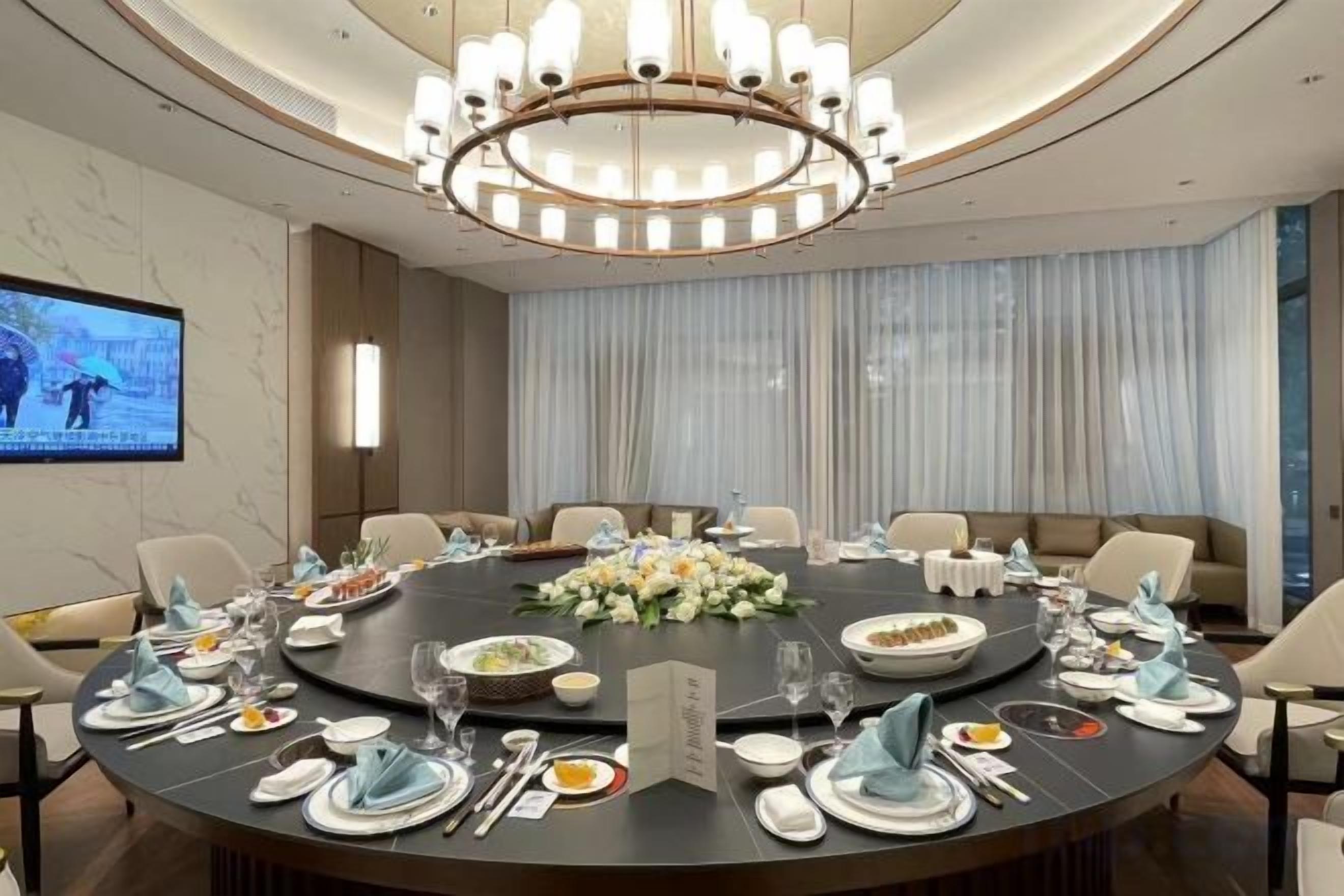 Une salle de conférence avec une table ronde préparée pour une réunion, un grand lustre et un écran diffusant une présentation.