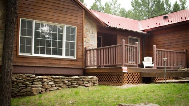 Une cabane en bois avec des fondations en pierre, une véranda dotée d’une balustrade et une chaise blanche.