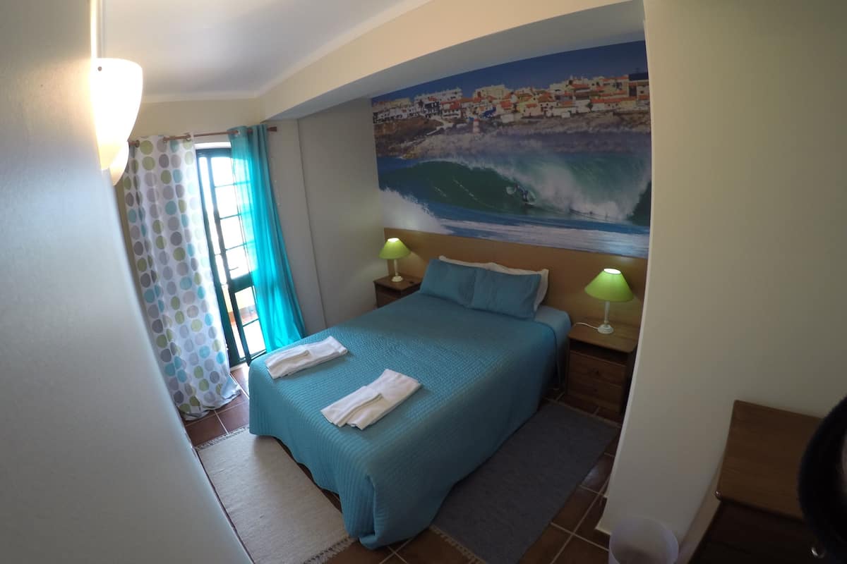 A bedroom with a bed, two bedside lamps, a large mural of a coastal scene, and a window with curtains.