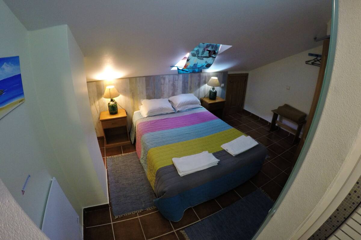A bedroom with a bed, two bedside tables with lamps, a small wooden stool, a wall-mounted TV, and a painting on the wall.