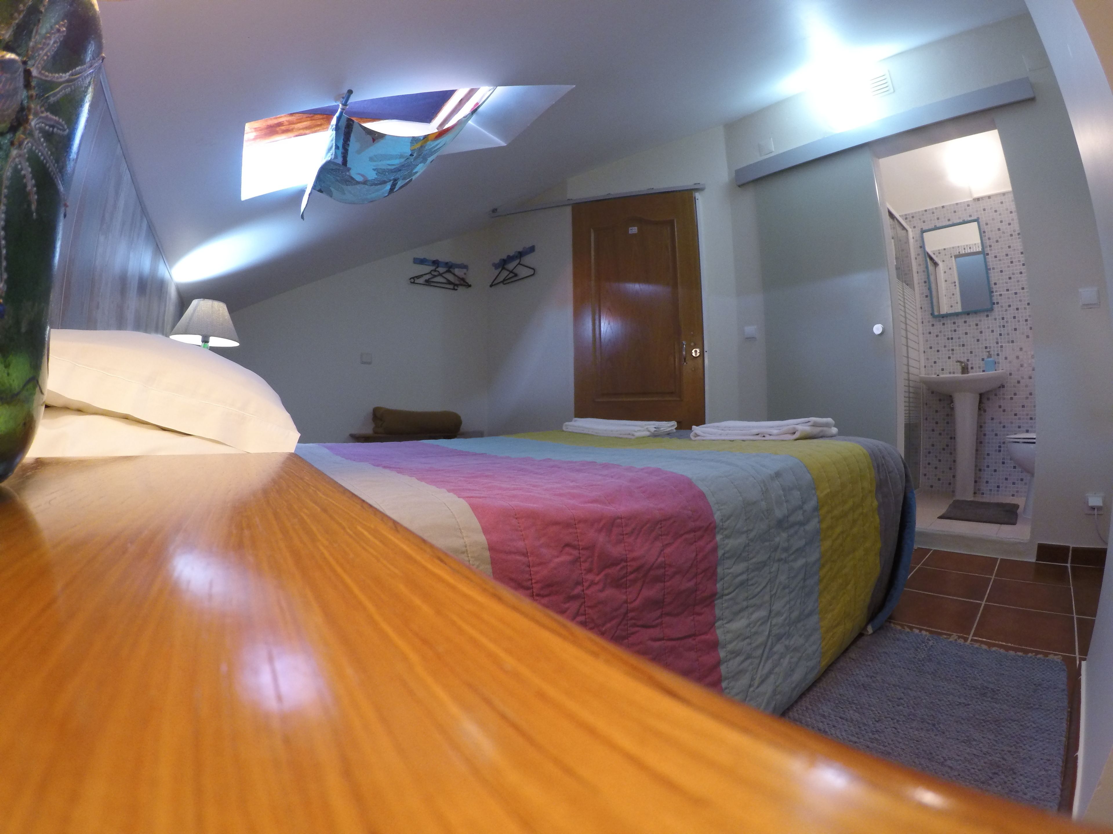 Une chambre avec un lit en bois, une couverture aux couleurs vives, une porte en bois et une salle de bain équipée d’un lavabo et d’un miroir.