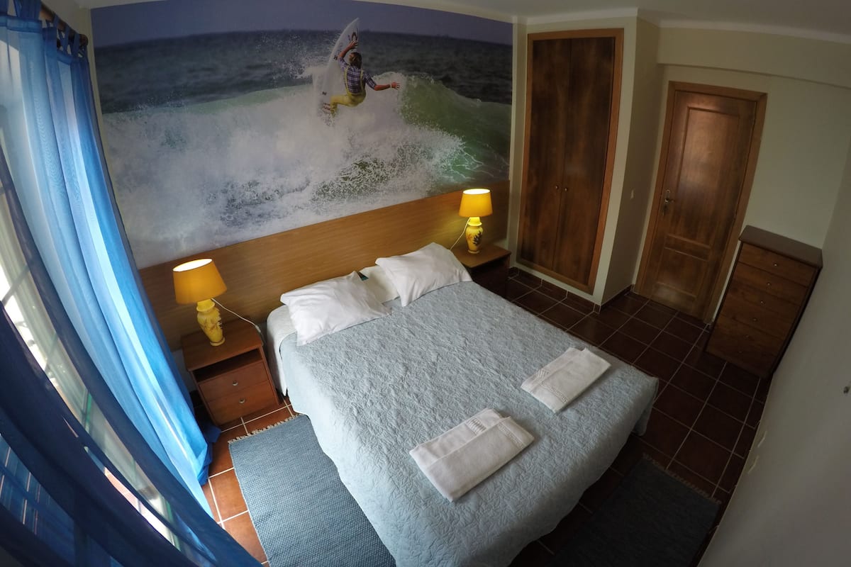 A bedroom with a bed, two bedside lamps, a wooden wardrobe, and a mural of a surfer on a wave.