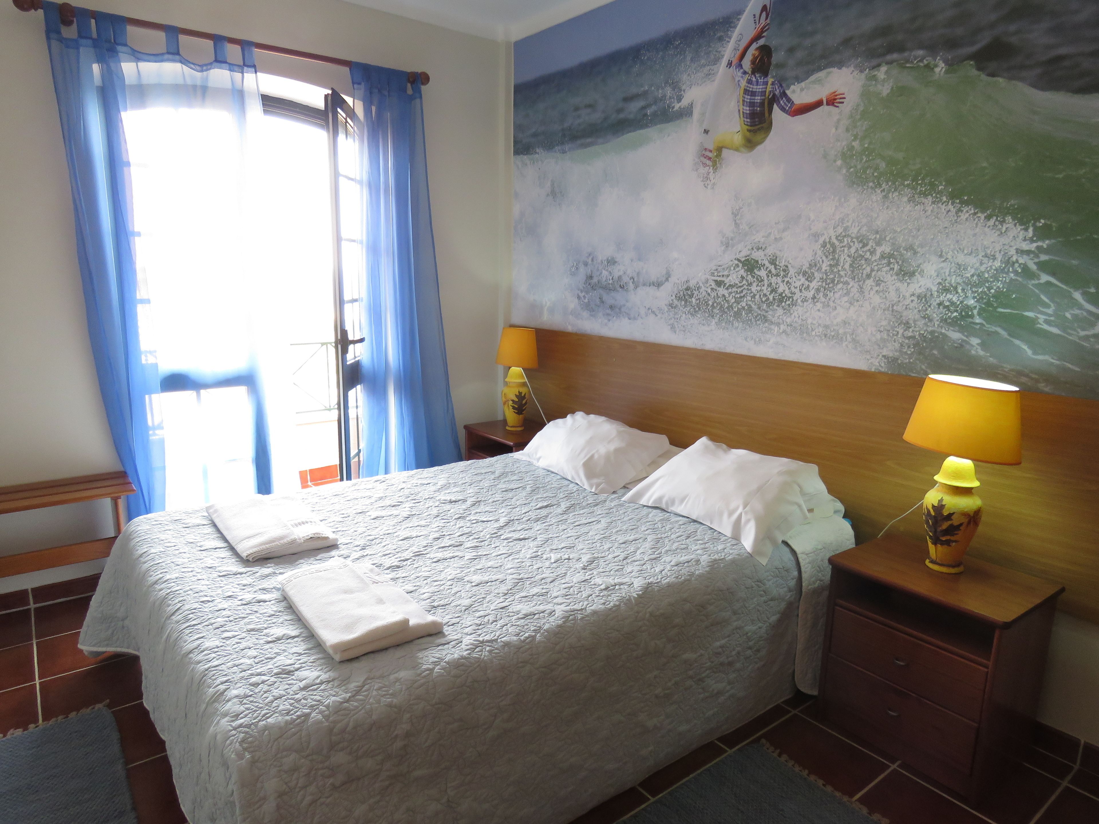 Una habitación de hotel con una cama, dos lámparas de noche, un ventanal con cortinas azules y un cuadro de un surfista sobre una ola.