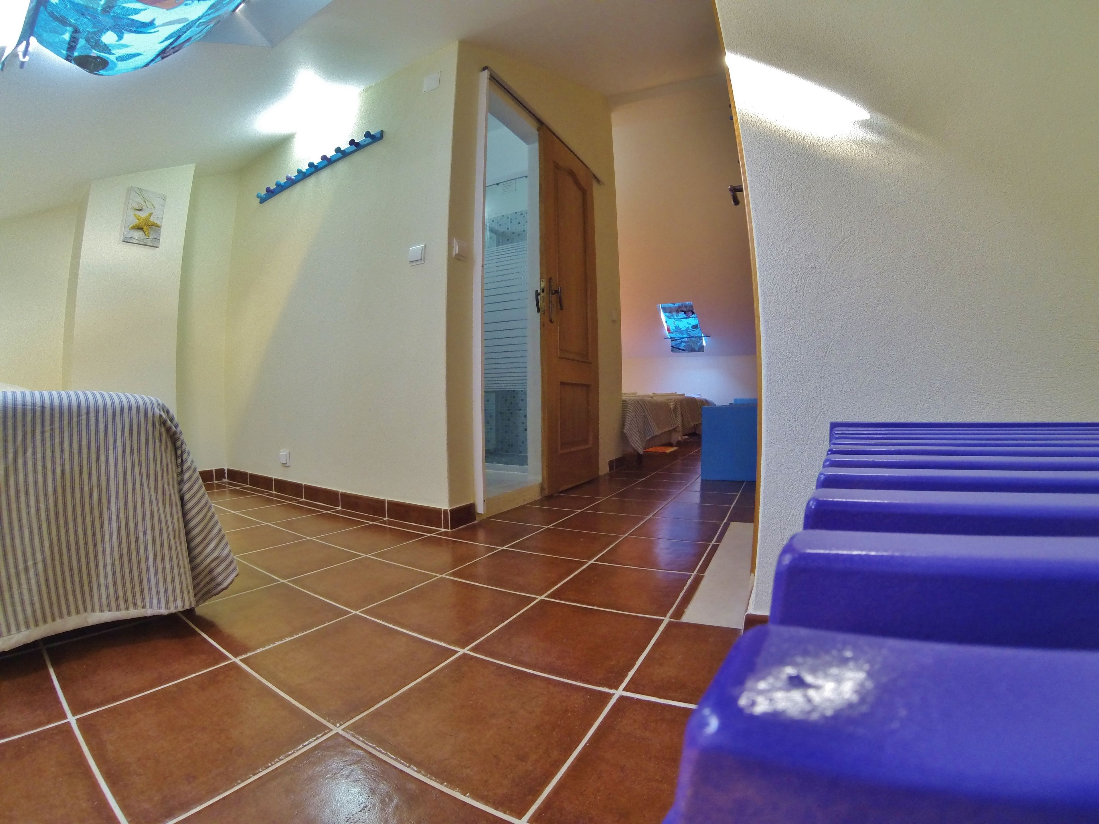 Una habitación de hotel con piso de baldosas, una cama con cubierta a rayas, una puerta de madera y un ventanal pequeño con persianas.