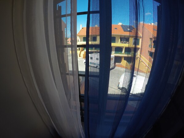 A view through a window with sheer curtains, revealing a building with a red roof and yellow walls.