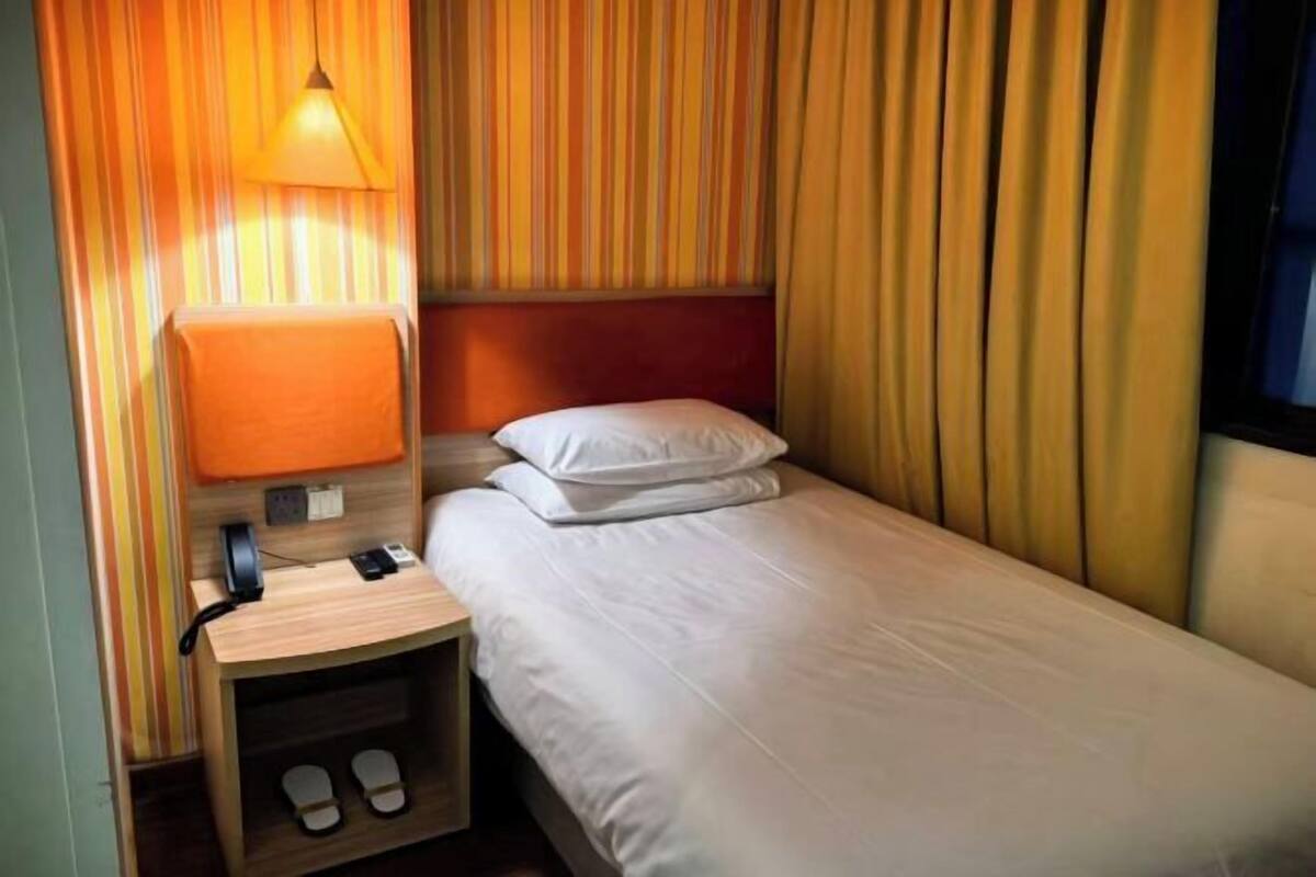 A single bed with white bedding, a bedside table with a lamp, and a pair of slippers.