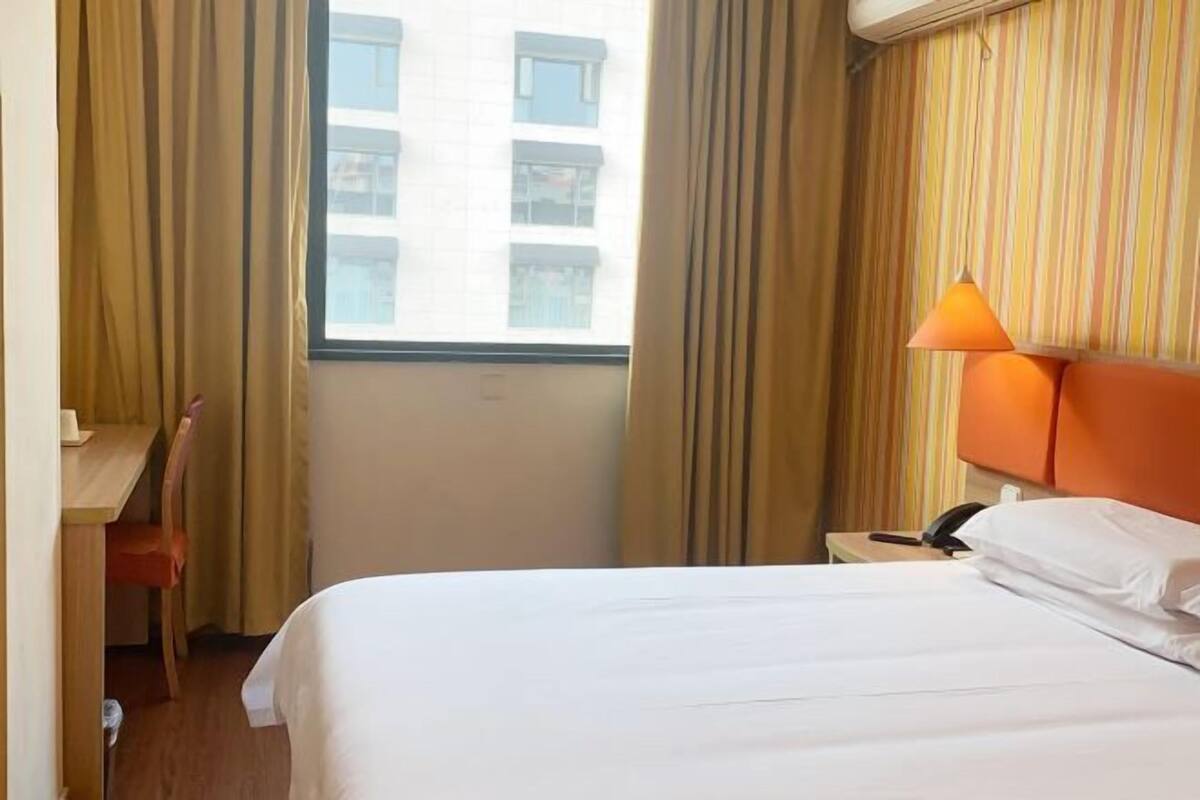 A hotel room with a bed, a desk, a chair, a nightstand with a lamp, and a window with curtains.