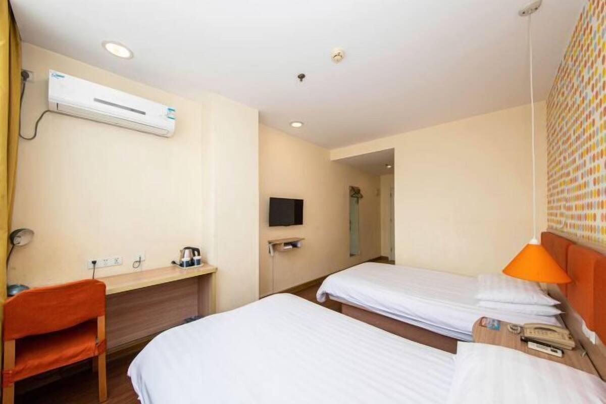 A hotel room with two beds, a desk, a chair, an air conditioning unit, a television, and a bathroom visible through an open door.