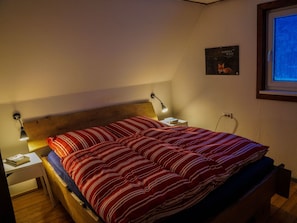 Una cama individual con cabecera de madera, manta a rayas rojas y blancas, lámpara de noche y una mesita con un libro.