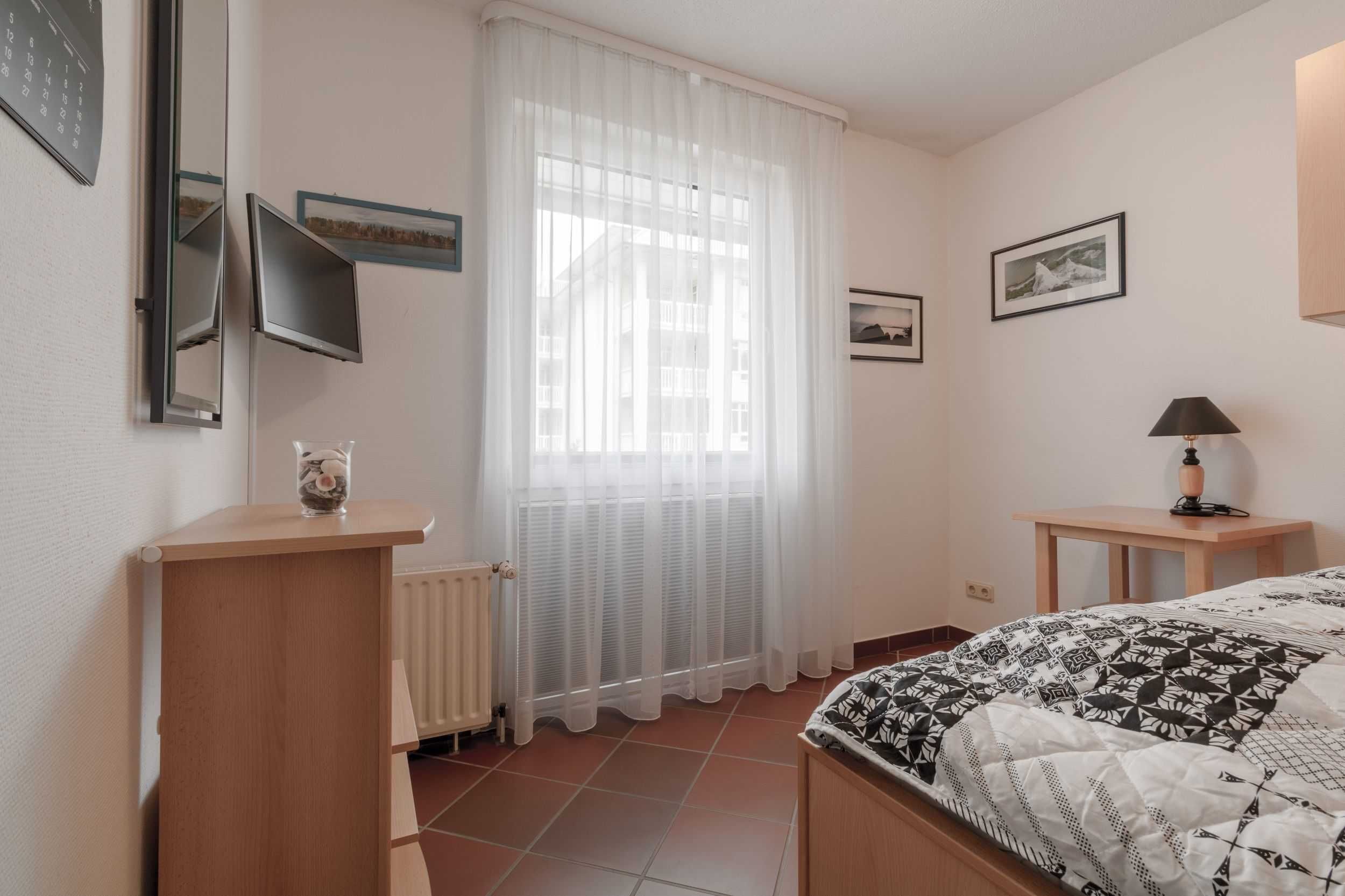 Une chambre à coucher avec un lit, une table de chevet avec une lampe, un radiateur, une fenêtre avec des rideaux légers et des tableaux encadrés accrochés au mur.