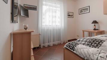 Une chambre à coucher avec un lit, une table de chevet avec une lampe, un radiateur, une fenêtre avec des rideaux légers et des tableaux encadrés accrochés au mur.