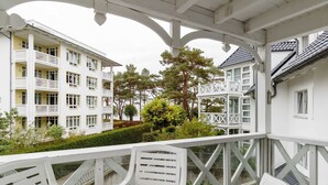 Ein weißer Balkon mit weißen Stühlen und einem Tisch, mit Blick auf ein Gebäude mit mehreren Balkonen und einen Garten mit Bäumen.