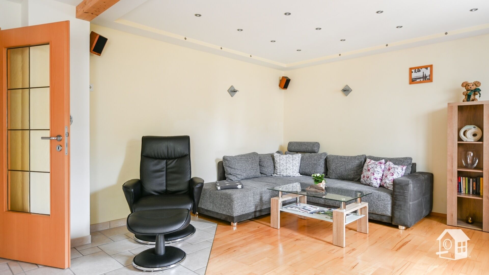 A living room with a sofa, a black leather chair, a glass coffee table, and a wooden bookshelf.