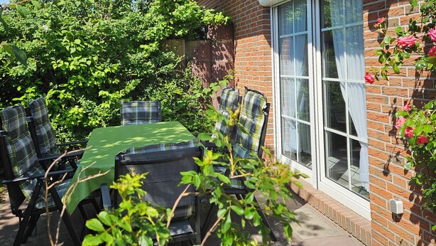 Eine Terrasse im Freien mit Tisch und Stühlen, umgeben von Grünpflanzen und einem Backsteinhaus.