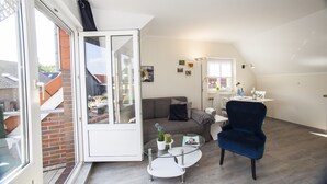 Uma sala de estar moderna com uma porta de vidro que dá para uma varanda, uma mesa de centro redonda de vidro, uma poltrona azul escura e um espaço de jantar com uma mesa e cadeiras brancas.