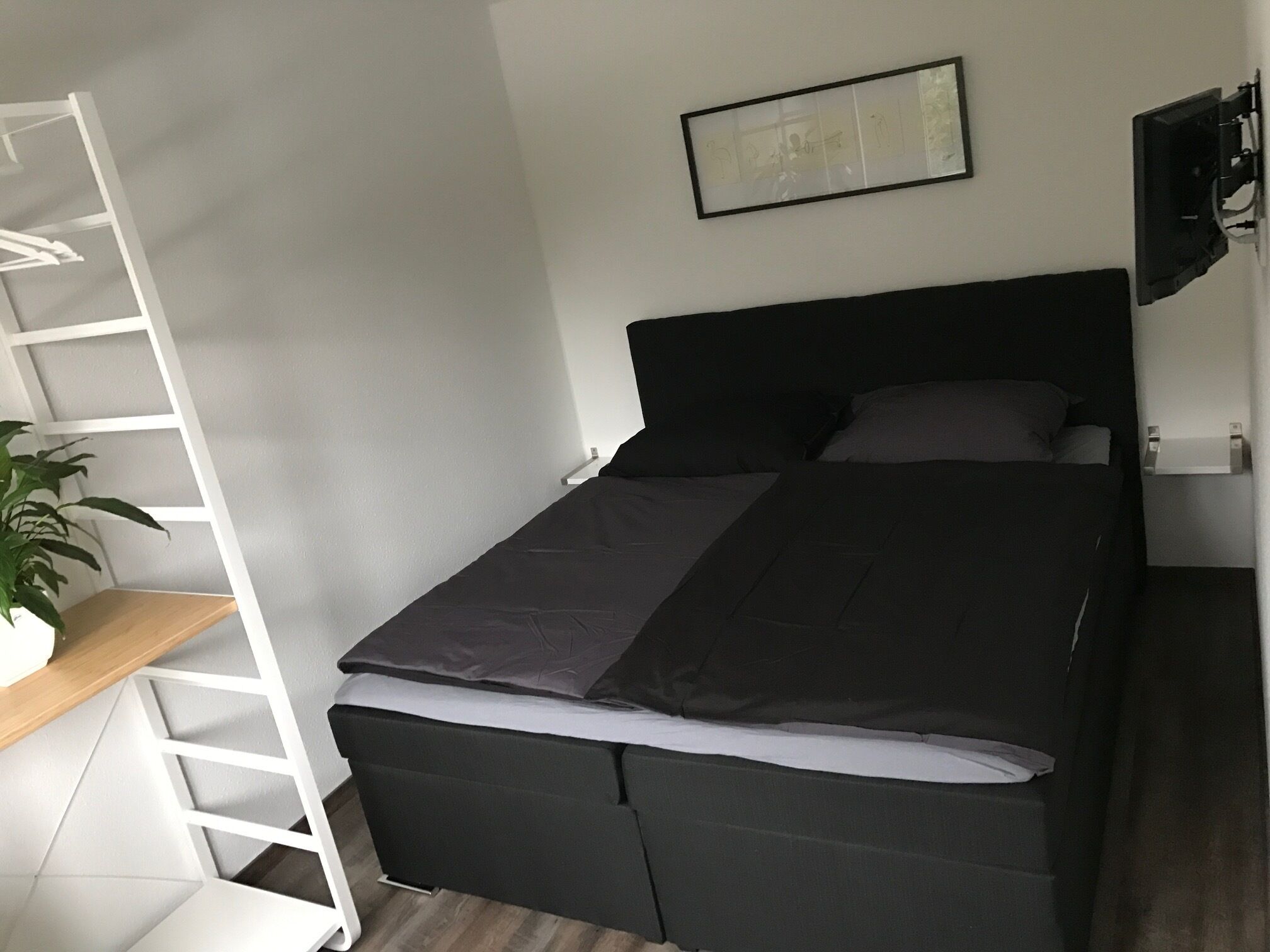 A compact bedroom with a bed, a white ladder-style shelf, a small desk, and a potted plant.
