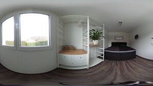 A bedroom with a curved window, a bed, a white shelving unit, and a potted plant.
