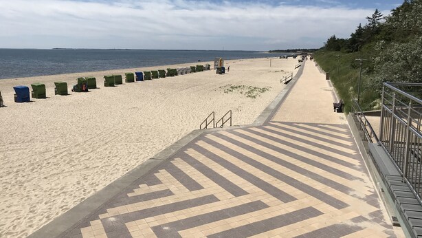 Una platja amb un passeig pavimentat que condueix al mar, amb gandules i una torre de socorristes.