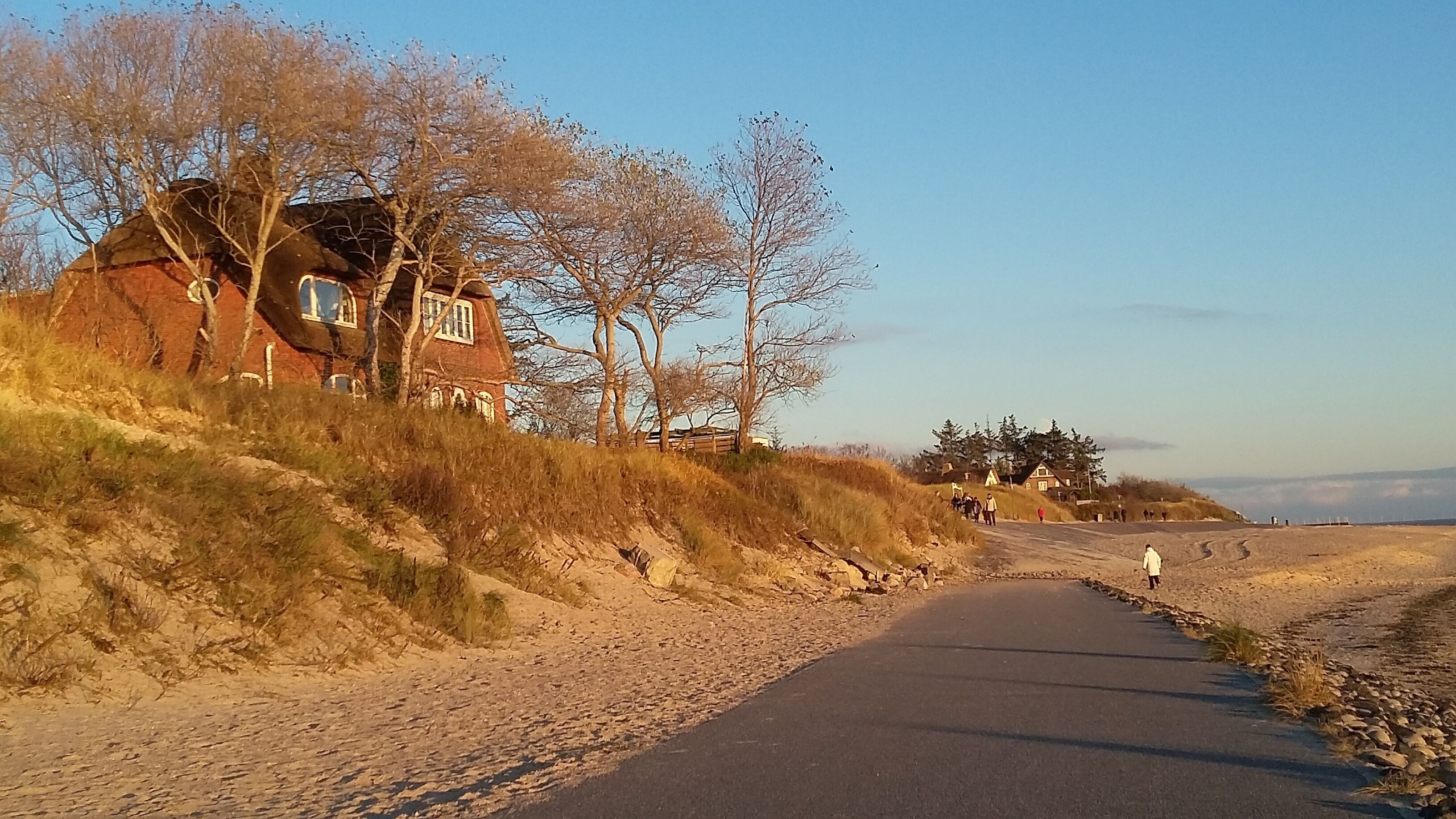 A coastal road with a paved path, a red brick house, and a group of people walking.