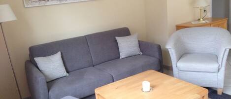 Una sala de estar con un sofá gris, un sillón gris, una mesa de centro de madera y un cuadro de una escena de playa en la pared.