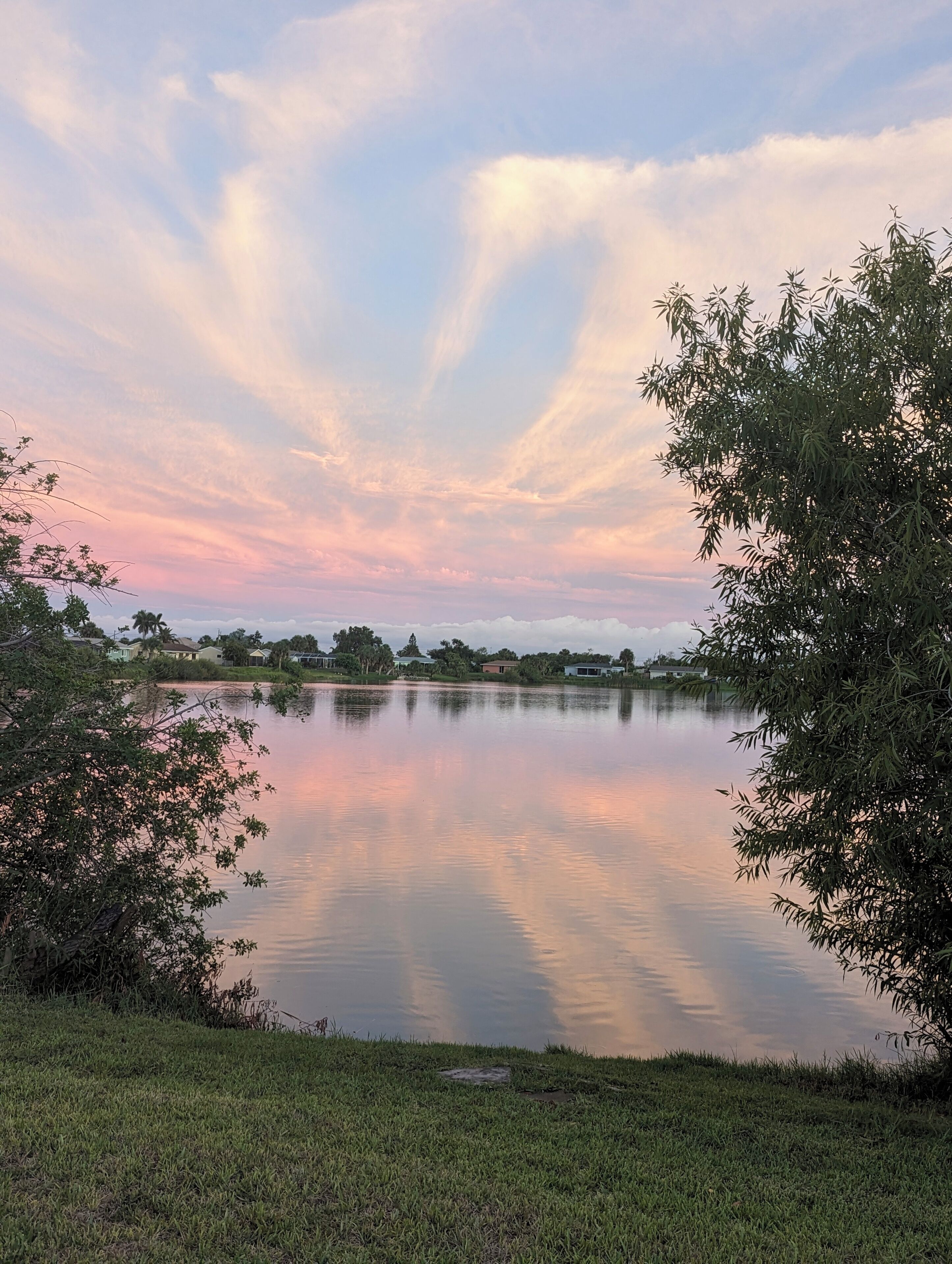 A tranquil lake with a clear reflection, surrounded by greenery and houses in the distance.