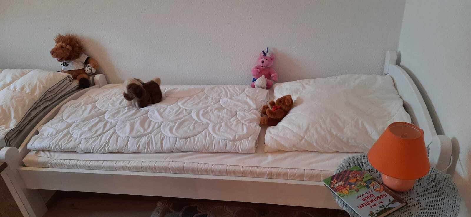 A bed with a white mattress and pillows, a teddy bear, a pink stuffed animal, and a book on a bedside table.
