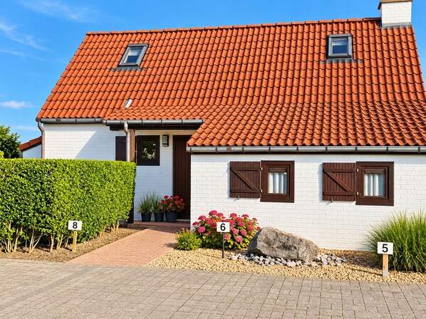 A two-story house with a red-tiled roof, white brick walls, and brown shutters. There are numbered signs indicating the house number and a small garden area with plants and flowers.
