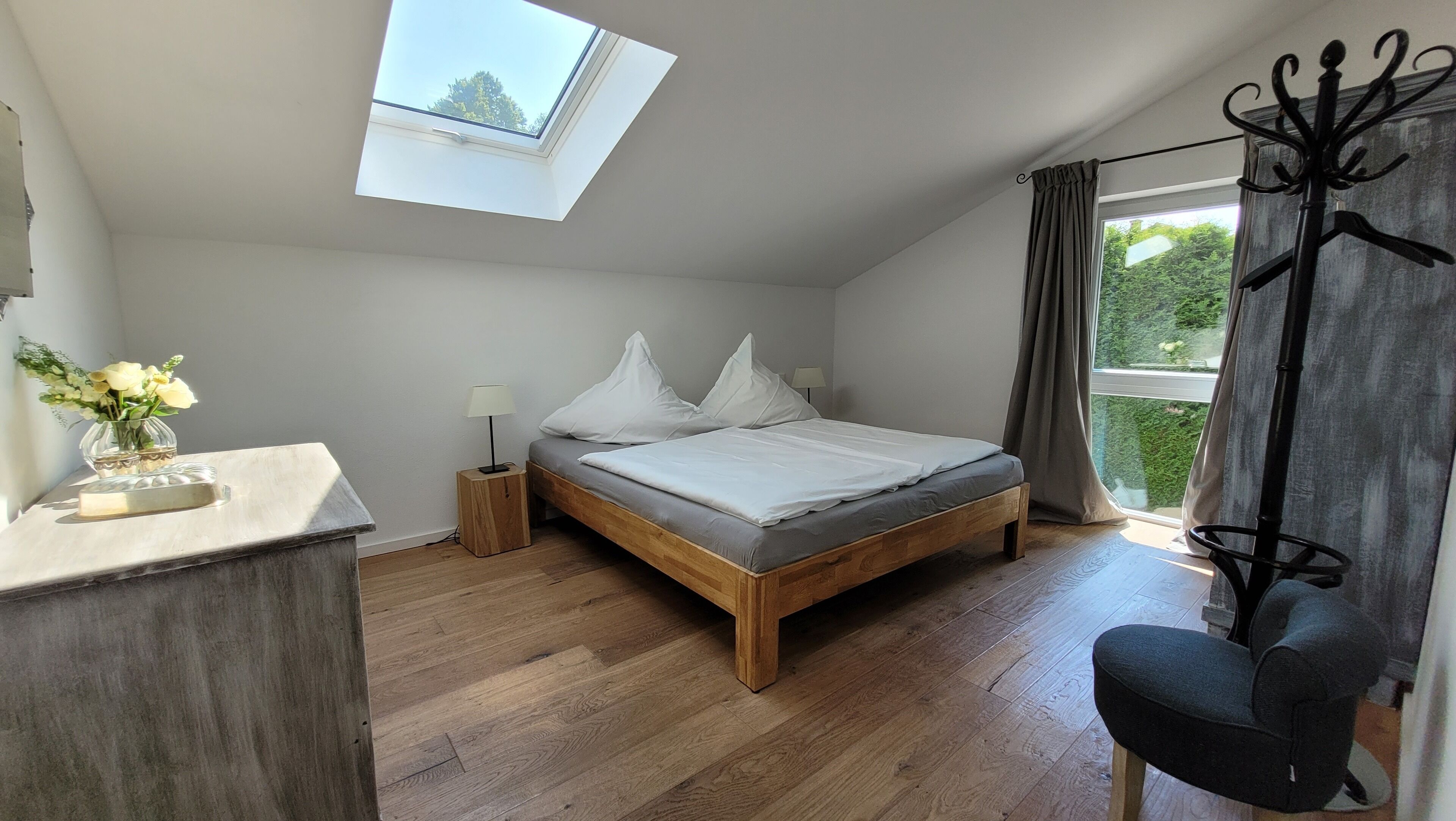Un dormitorio con tragaluz, una cama con ropa de cama blanca, una mesita de noche con lámpara, una silla y una ventana con cortinas.
