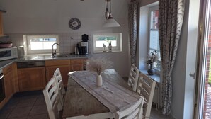 A kitchen with wooden cabinets, a dining table, chairs, a clock, a coffee maker, and a window with curtains. - Anti-allergy and comfortable vacation home (Dornum)