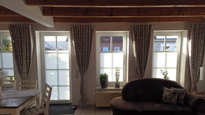 A living room with a sofa, a dining table with chairs, and a window with curtains. - Anti-allergy and comfortable vacation home (Dornum)