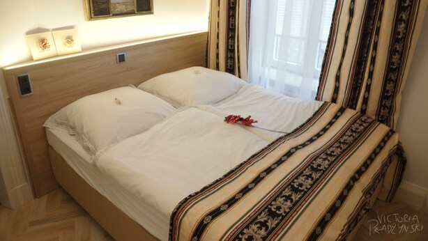 Una cama bien hecha con edredón blanco y almohada decorativa, cabecera de madera y mesita de noche con lámpara.