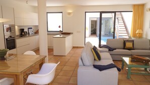 Un salon moderne avec un canapé, une table basse en verre et un coin repas comprenant une table et des chaises. On y trouve également une cuisine équipée avec des placards et des appareils électroménagers, et une vue sur un balcon avec une piscine.
