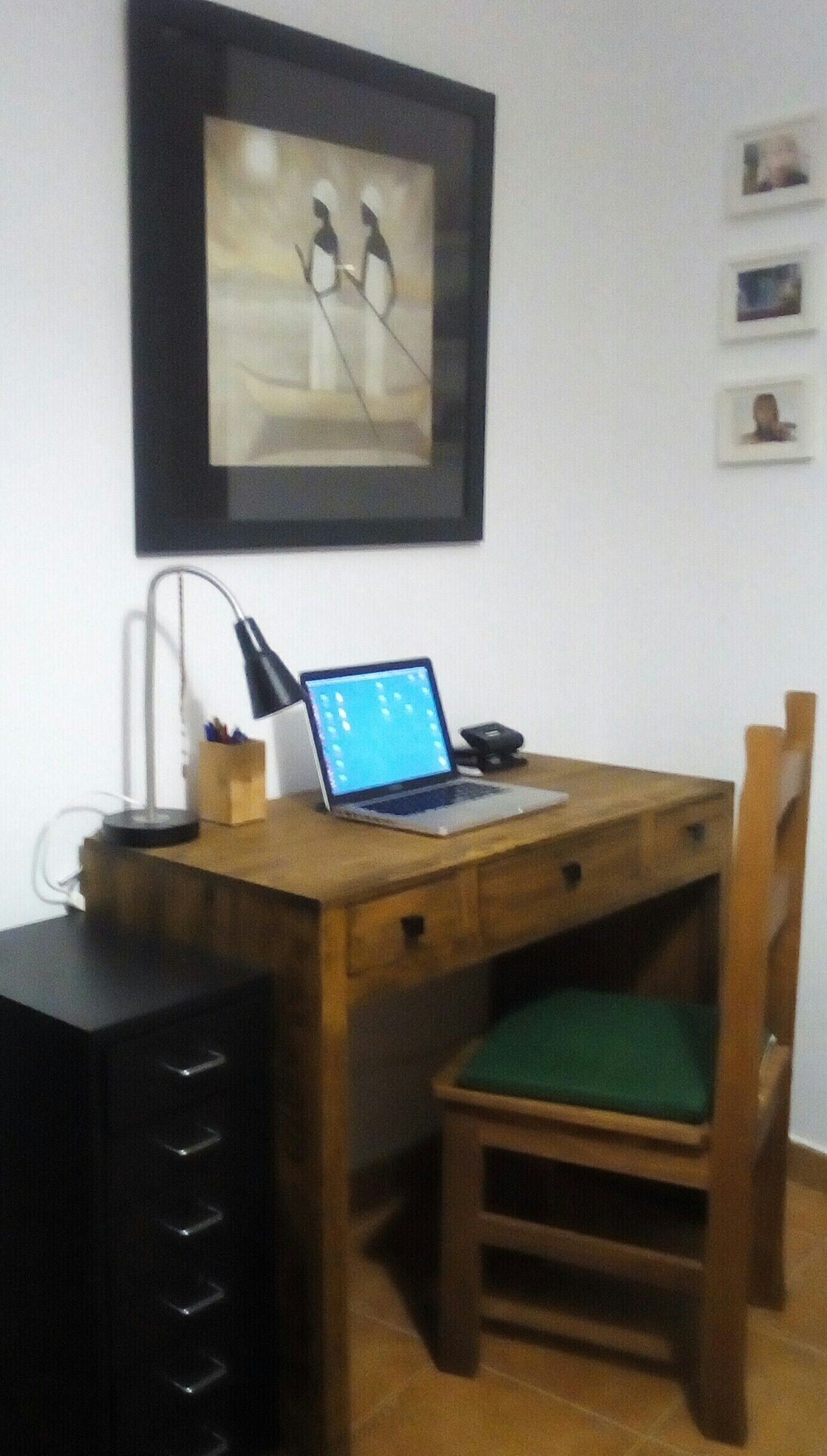 A wooden desk with a laptop, a lamp, and a chair with a green cushion. There are framed pictures on the wall and a black filing cabinet to the left.