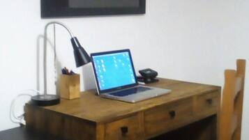 A wooden desk with a laptop, a lamp, and a chair with a green cushion. There are framed pictures on the wall and a black filing cabinet to the left.