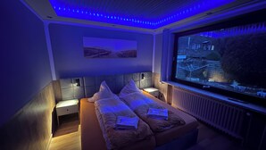 Un dormitorio con cama, mesita de noche, televisor montado en la pared y ventana con vista a una casa.